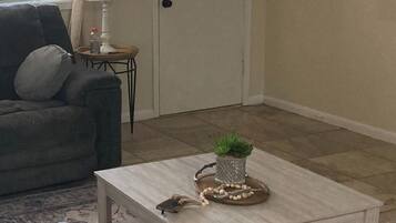 A living room with a grey sofa, a white ottoman, a wooden coffee table with a plant and decorative items, a lamp, and a door with a sign.
