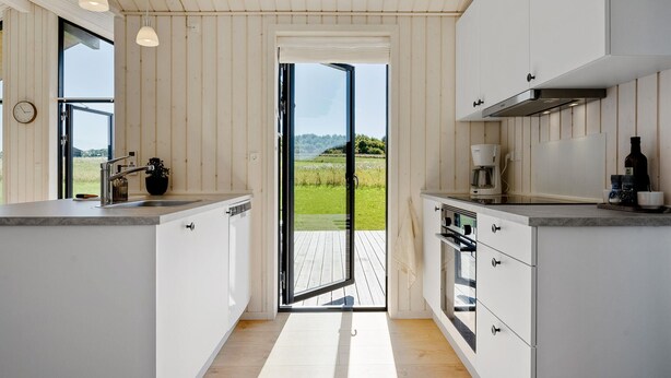 Une cuisine moderne avec des armoires blanches, un four encastré, une machine à café et une vue sur un paysage verdoyant par une porte vitrée.