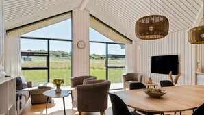 Une pièce spacieuse avec une grande table en bois, des sièges confortables, un téléviseur et une vue sur un champ verdoyant grâce à de grandes fenêtres.