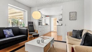 Una sala de estar moderna con sofá, sillón y mesa de centro. Se observa una cocina al fondo con mesa y sillas.