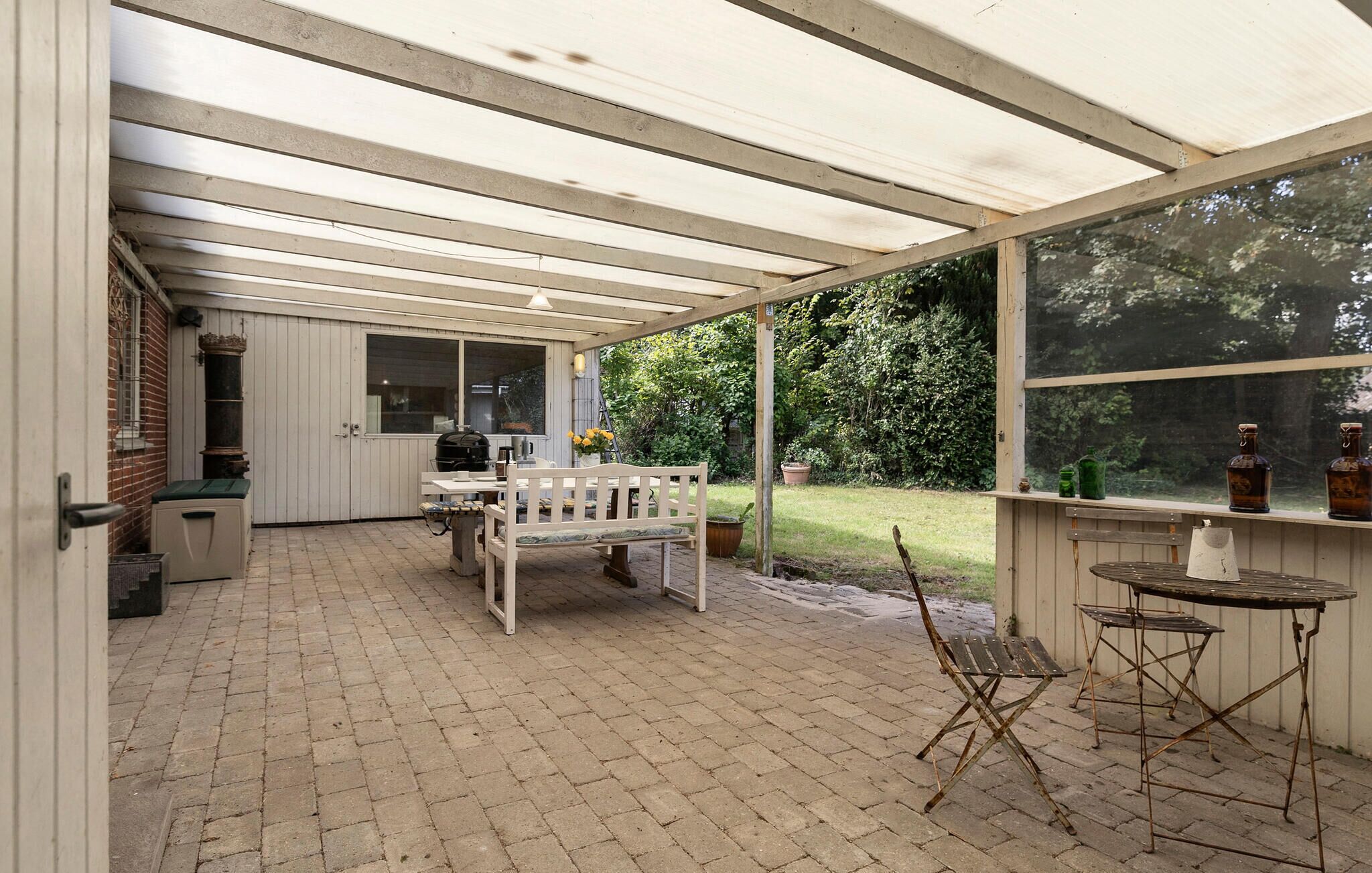 A covered patio area with a brick floor, a wooden bench, a table with chairs, and a view of the garden.