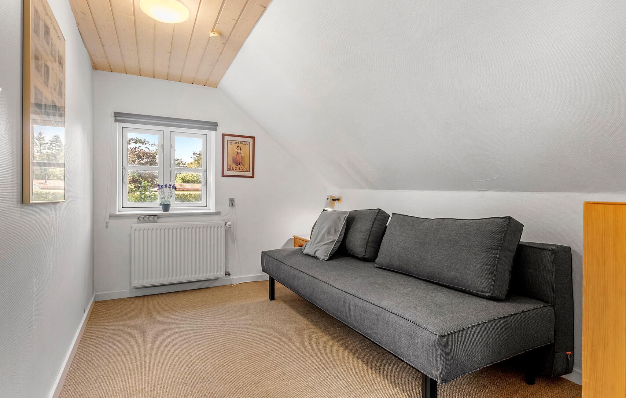 A cozy room with a grey sofa, a window with a view of greenery, a radiator, and a wooden ceiling.