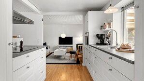 Una cocina moderna con armarios blancos, encimera negra y suelo de madera. Hay una zona de comedor con una mesa y sillas, y un televisor montado en la pared.