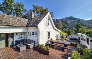 Une maison blanche avec un toit en pente, une terrasse avec des meubles d’extérieur et une vue sur les montagnes en arrière-plan.