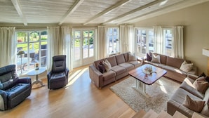 A spacious living room with a large sectional sofa, a coffee table, and a fireplace. There are multiple windows providing natural light and views of the outdoors.
