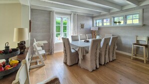 A dining area with a round table and six chairs, a high ceiling with exposed beams, and a view of greenery outside.