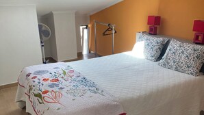 Une chambre avec une lucarne, un lit recouvert d’une courtepointe à motifs floraux, deux coussins à motifs et une lampe rouge posée sur une table de chevet.