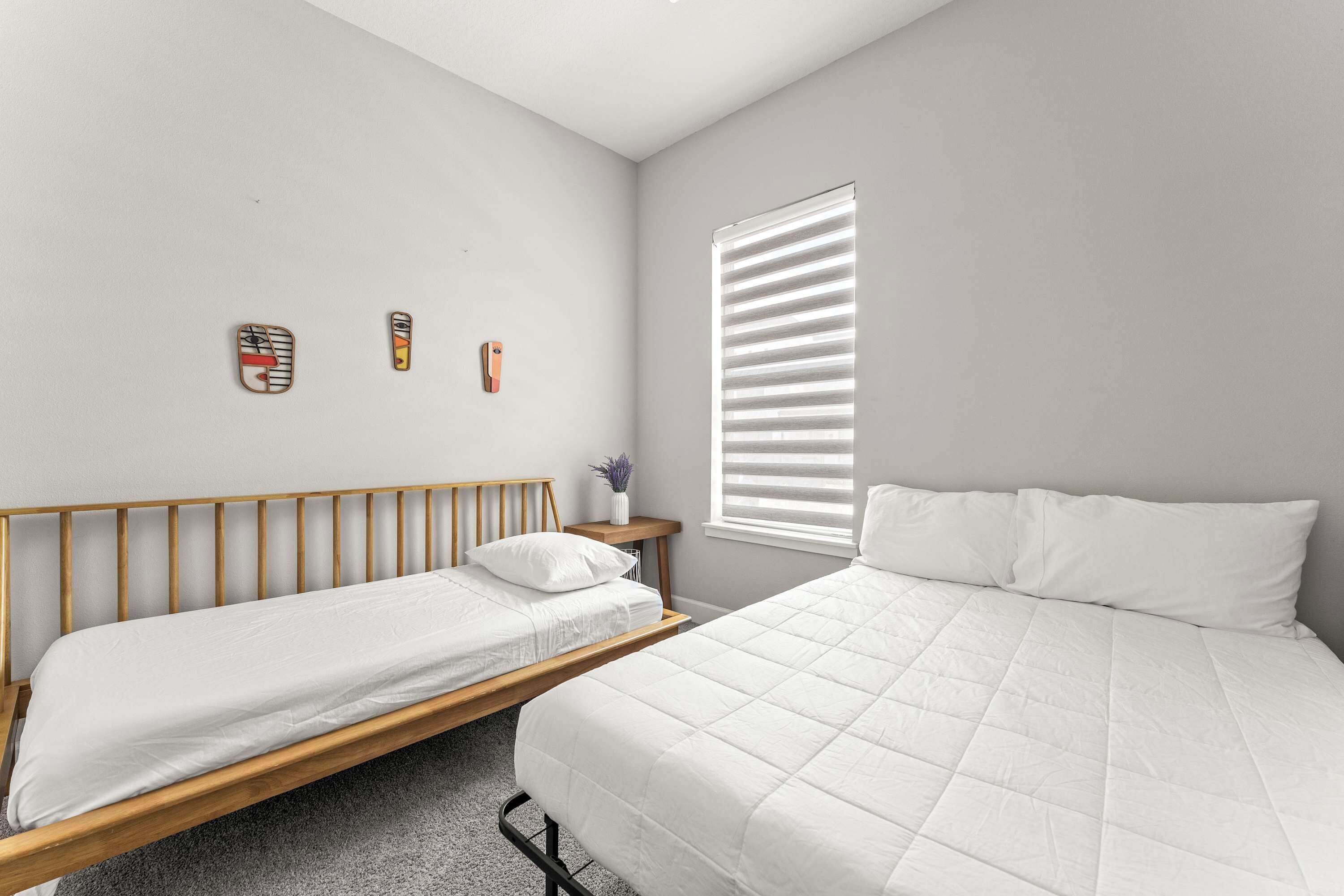 A bedroom with two beds, a wooden headboard, a small bedside table with a vase, and a window with blinds.