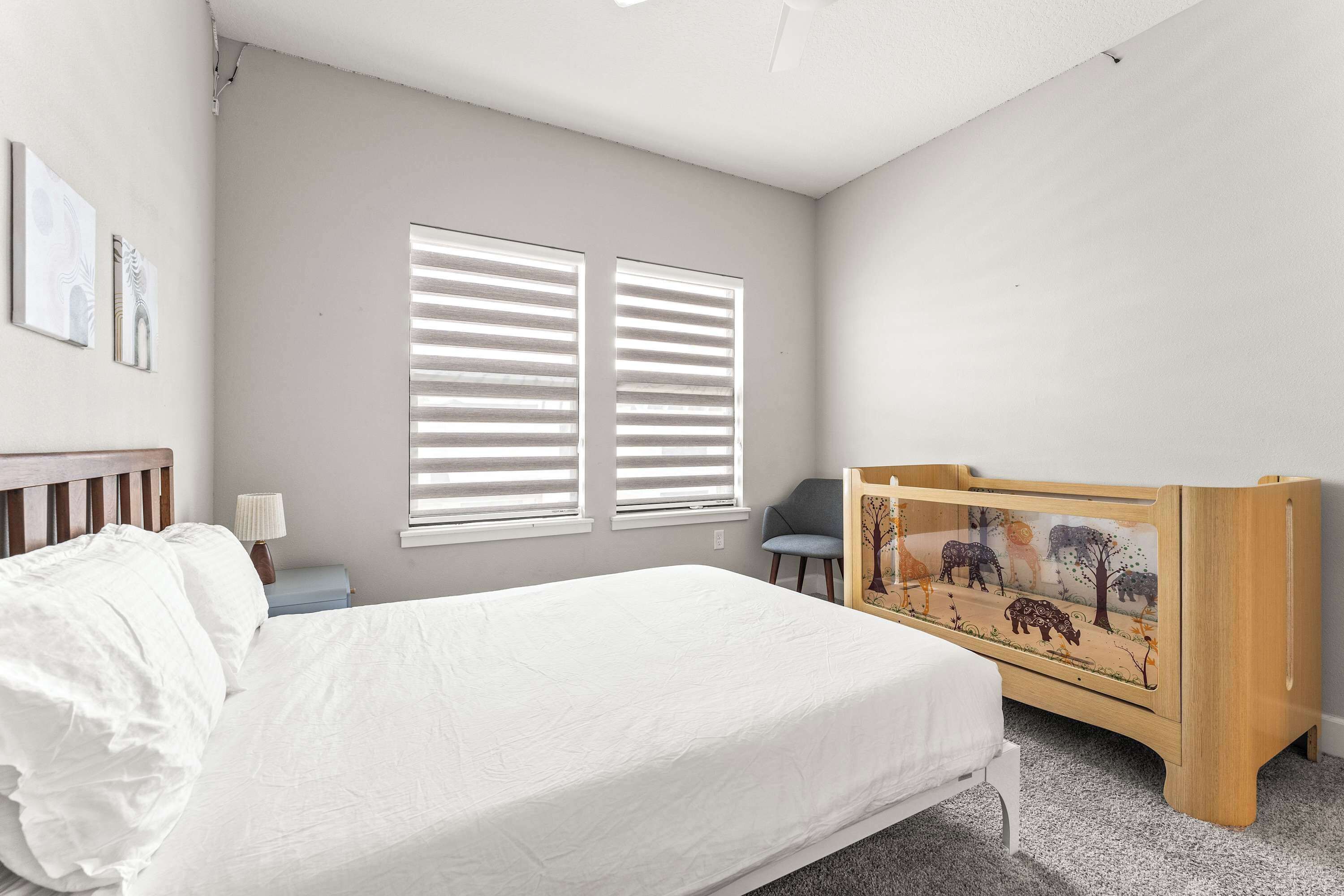 A bedroom with a bed, two windows with blinds, a chair, a crib with a forest mural, and a lamp on a nightstand.