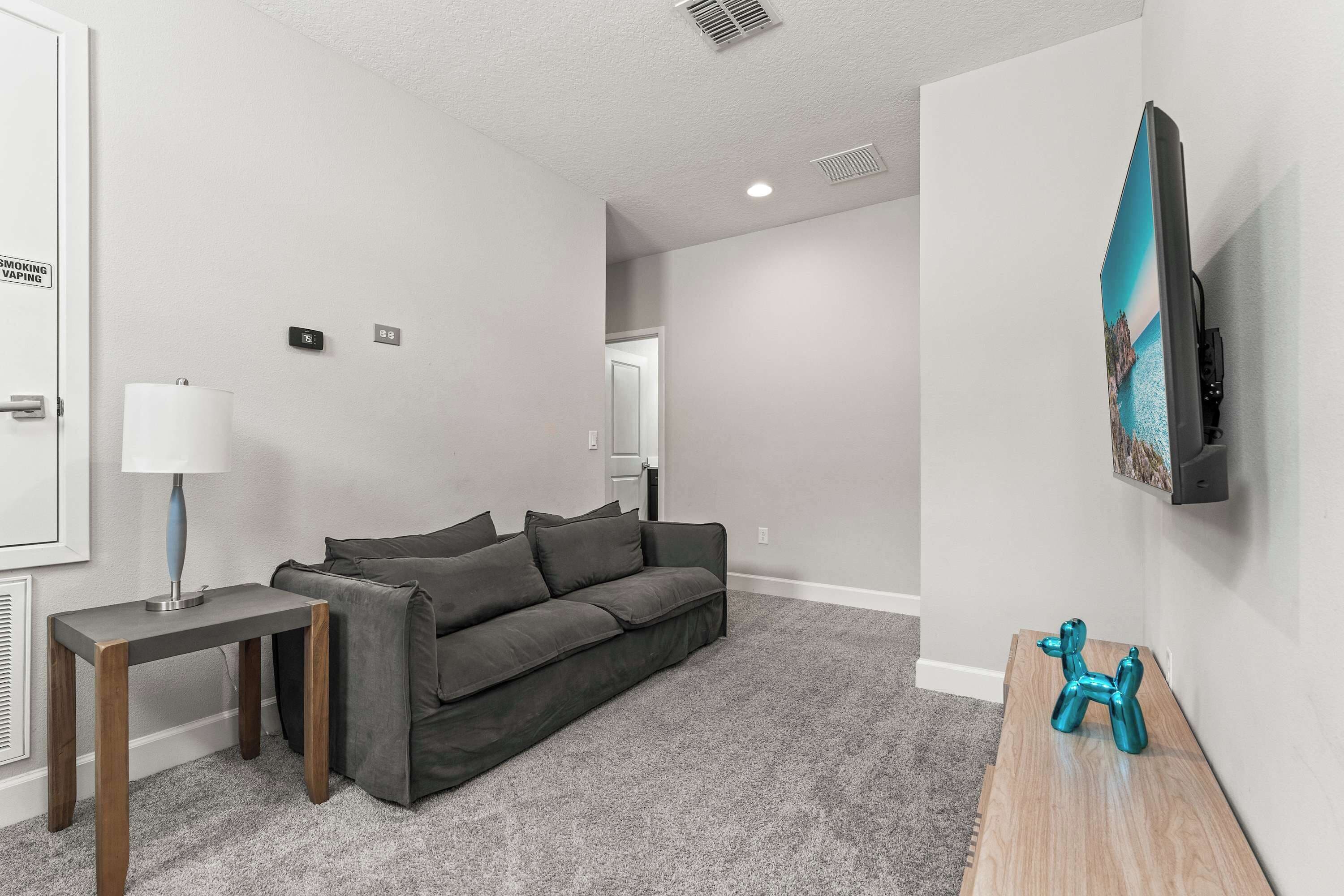 A modern living room with a grey sofa, a small side table with a lamp, a mounted TV, and a wall-mounted sculpture.