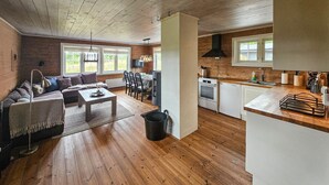 Un vaste espace de vie doté d’un plancher en bois, d’un canapé, d’une table basse et d’une table à manger avec des chaises. On aperçoit une cuisine équipée d’appareils modernes et une fenêtre à l’arrière-plan.