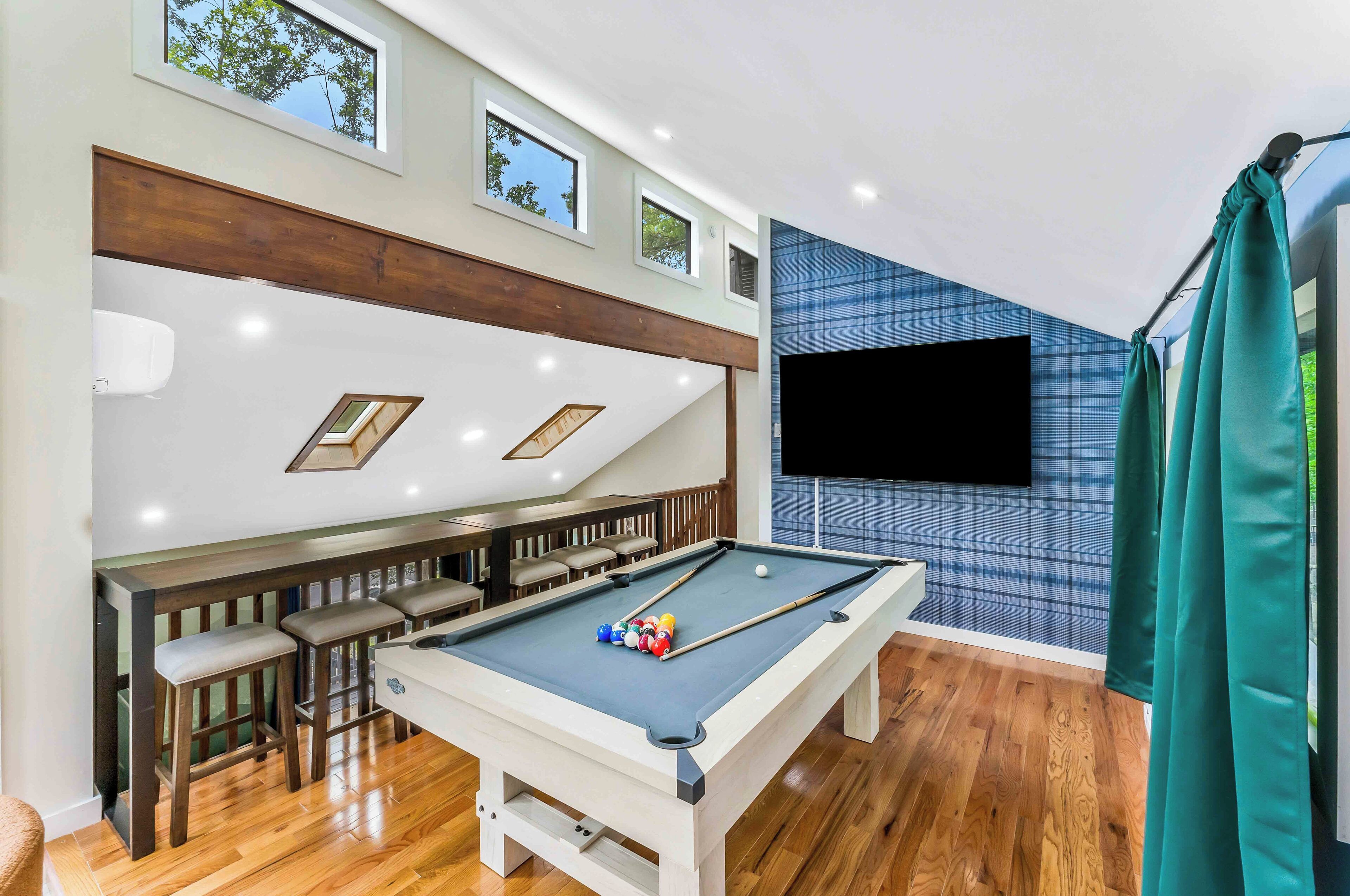 Una mesa de billar con taco, un área de bar con taburetes y un televisor montado en la pared.