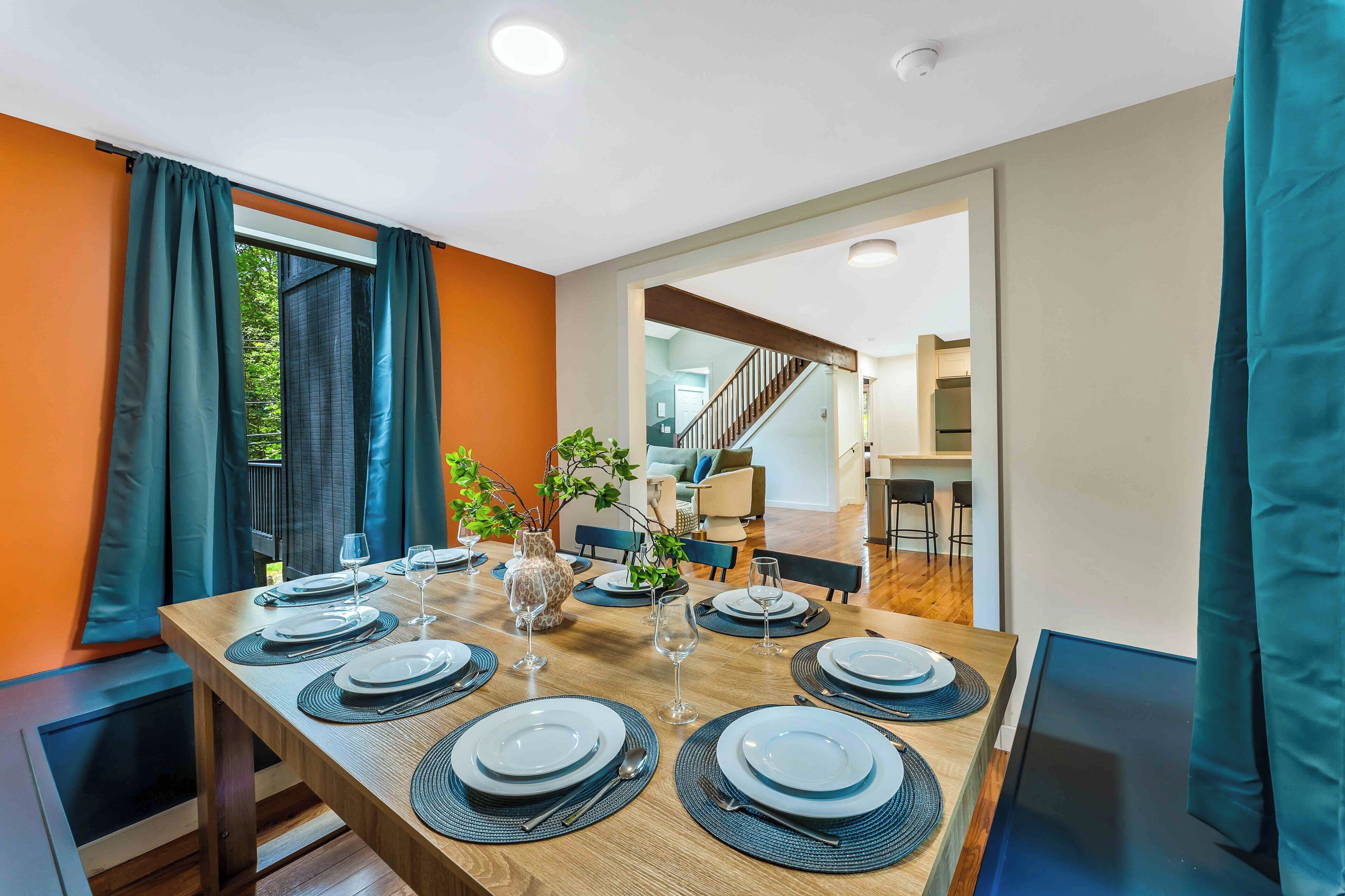 A dining area with a wooden table set for six, a staircase in the background, and a view of the outdoors.