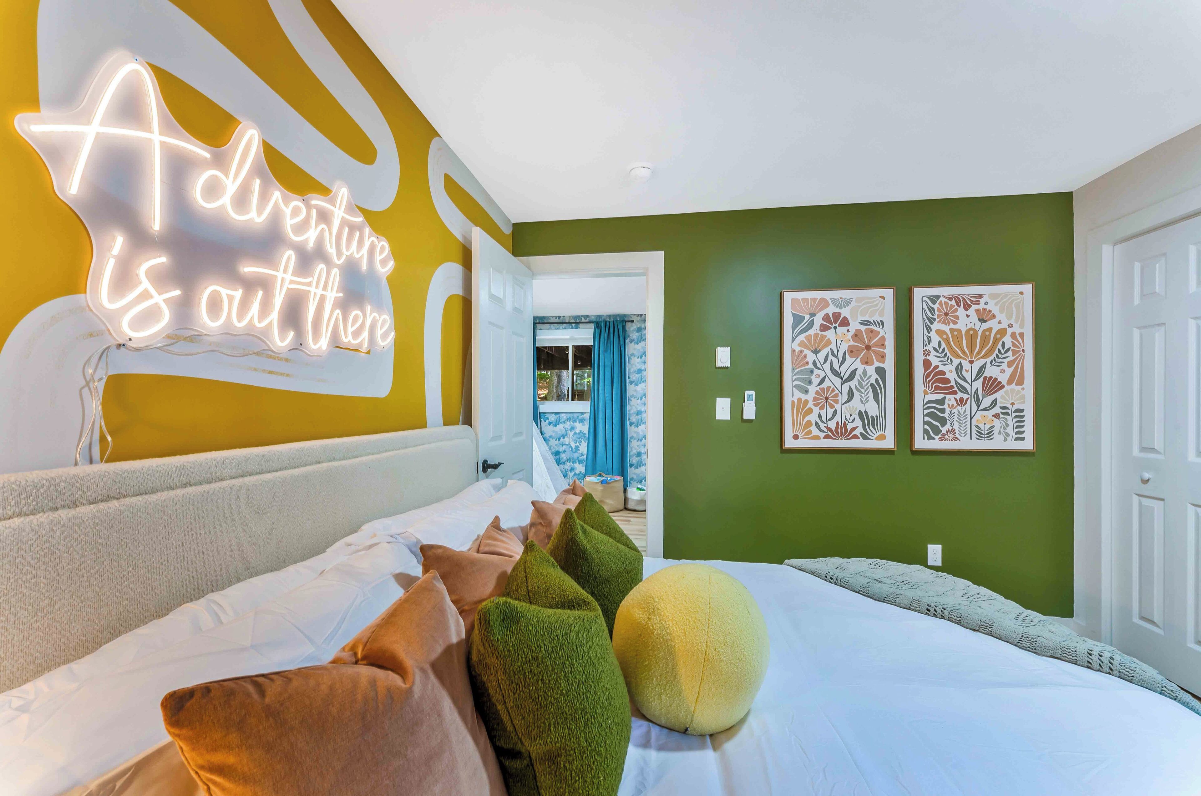 Une chambre d’hôtel avec un lit, des oreillers, un panneau lumineux au néon et deux œuvres d’art à motifs floraux accrochées au mur.