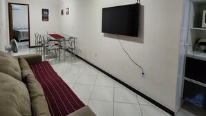 Un salon comprenant un canapé, un téléviseur fixé au mur, une table à manger avec des chaises et un sol carrelé.