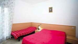 Une petite pièce avec deux lits, l’un recouvert d’une couverture rouge et l’autre d’une couverture à carreaux. On y trouve une petite table avec une lampe, et un tableau accroché au mur.