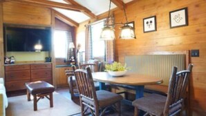 A wooden cabin interior with a dining table, chairs, a kitchen area, and a large window.
