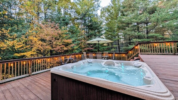 A hot tub on a wooden deck with a railing, surrounded by trees and outdoor furniture.