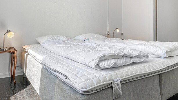 A neatly made bed with a white duvet and pillows, a bedside table with a lamp, and a grey upholstered bench. - Cozy apartment in Højer with WiFi (Højer)