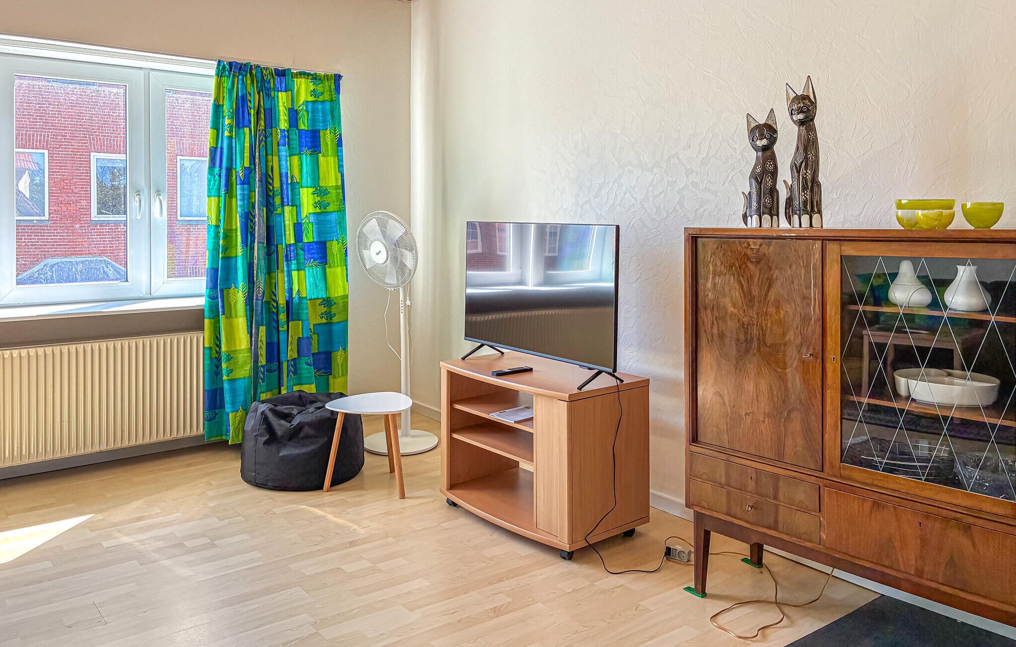 Une pièce avec un téléviseur posé sur un meuble en bois, un buffet orné d’objets décoratifs, un fauteuil en forme de sac noir et une fenêtre avec des rideaux.