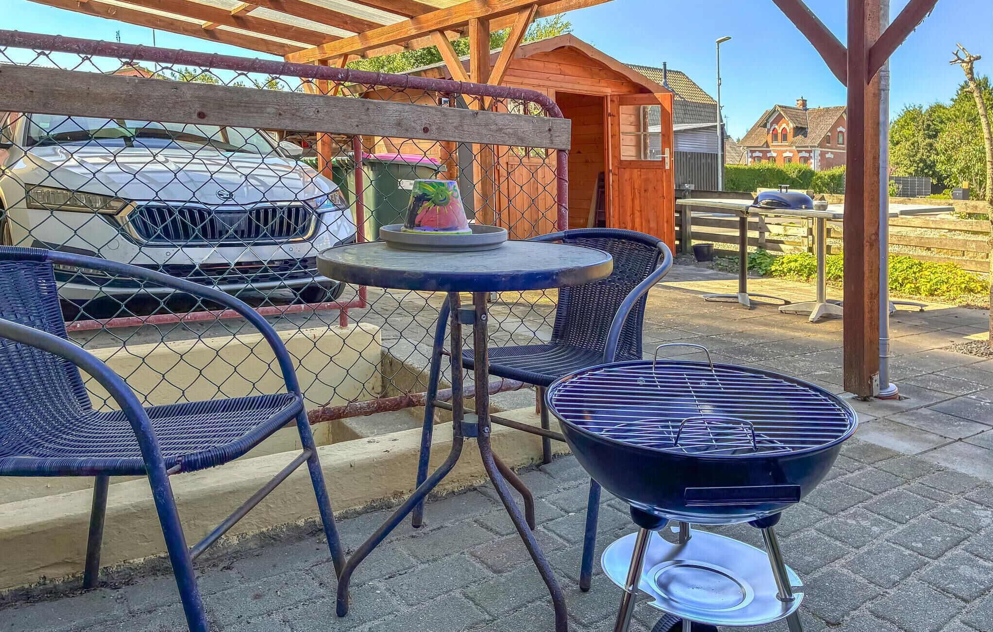 Een terras met een ronde tafel en twee stoelen, een barbecue en een auto achter een hek van gaas.
