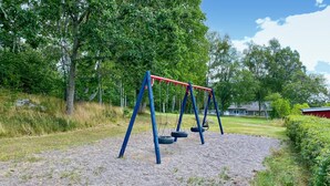 Ein Spielplatz mit Schaukeln und einer Reifenrutsche auf Kies, umgeben von Bäumen und einem Haus im Hintergrund.