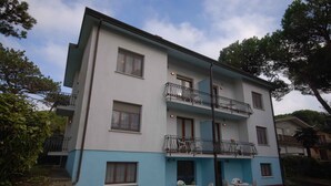Un edificio residencial de dos plantas con un balcón en el piso superior, una pequeña zona de estar al aire libre con mesas y sillas, y una entrada visible.