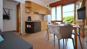 Een kleine keuken met houten kastjes, een eettafel met stoelen en een televisie op een standaard.