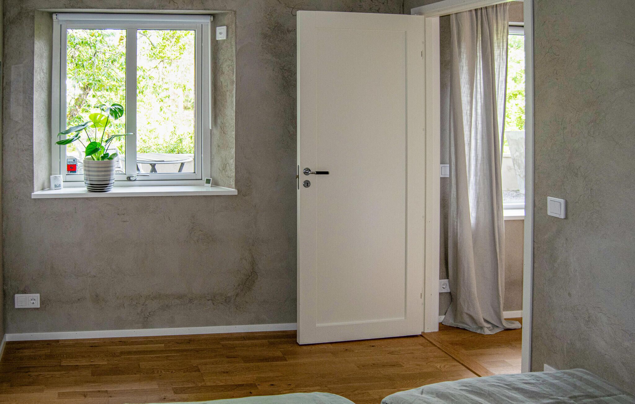Une pièce avec un plancher en bois, une porte blanche, une fenêtre avec une plante et une vue sur la verdure à l’extérieur.