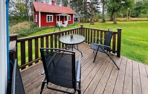 Une terrasse en bois avec une table ronde et deux chaises, d’où l’on peut admirer une maison rouge et une cour verdoyante.