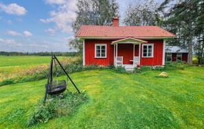 Une maison en bois rouge, dotée d’un porche, entourée d’un champ verdoyant et d’arbres.