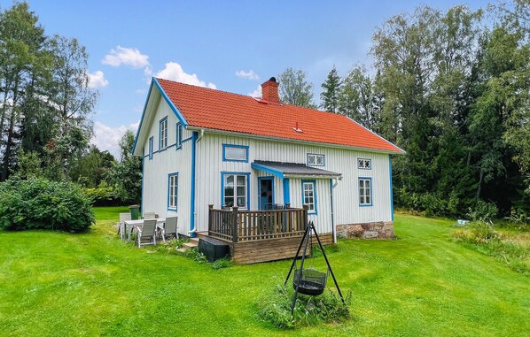 Une maison à deux étages avec un toit recouvert de tuiles rouges, des murs blancs et des bordures bleues. Elle possède une terrasse en bois avec des meubles d’extérieur et une balançoire.