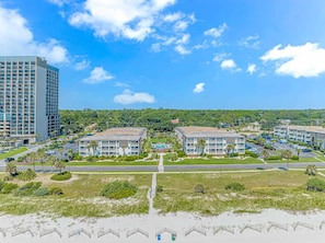A coastal hotel with multiple buildings, a parking lot, and a beach in the foreground.