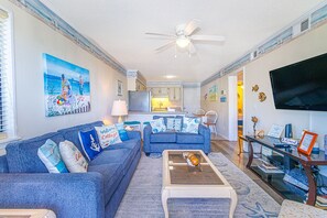 A living room with a sofa, coffee table, and a TV. There is a kitchen in the background.