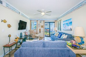 A living room with a ceiling fan, a TV, a sofa, a coffee table, and a view through a glass door to the outside. - Ocean Forest Villas C-114 (Myrtle Beach)