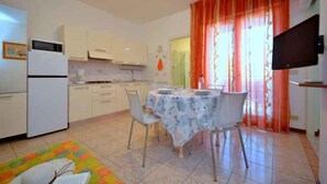 A compact kitchen with white cabinets, a dining table with chairs, a refrigerator, and a TV mounted on the wall. - Holiday flat with air conditioning and balcony (Bibione)