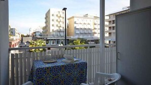 A balcony with a table covered in a patterned tablecloth, white chairs, and a view of surrounding buildings. - Holiday flat with air conditioning and balcony (Bibione)