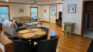 Una sala de estar acogedora con sofá, mesa de centro y sillones. Hay un televisor y una pequeña mesa con lámpara.