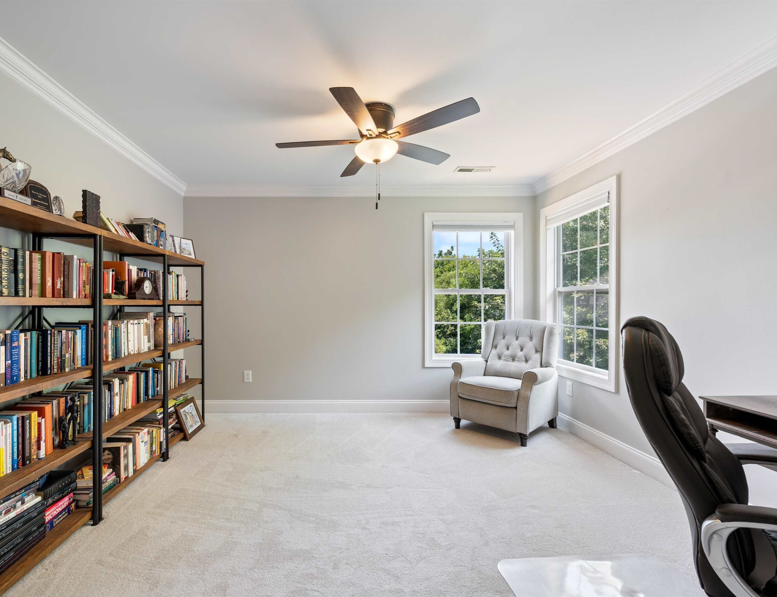 A spacious room with a ceiling fan, a large bookshelf filled with books, a comfortable armchair, and a view through a large window.