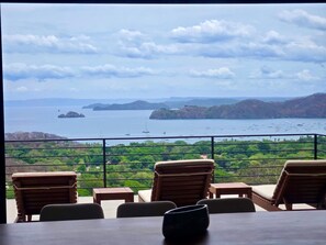 Terraza con sillas y mesas de madera con vista a una bahía con barcos y vegetación.