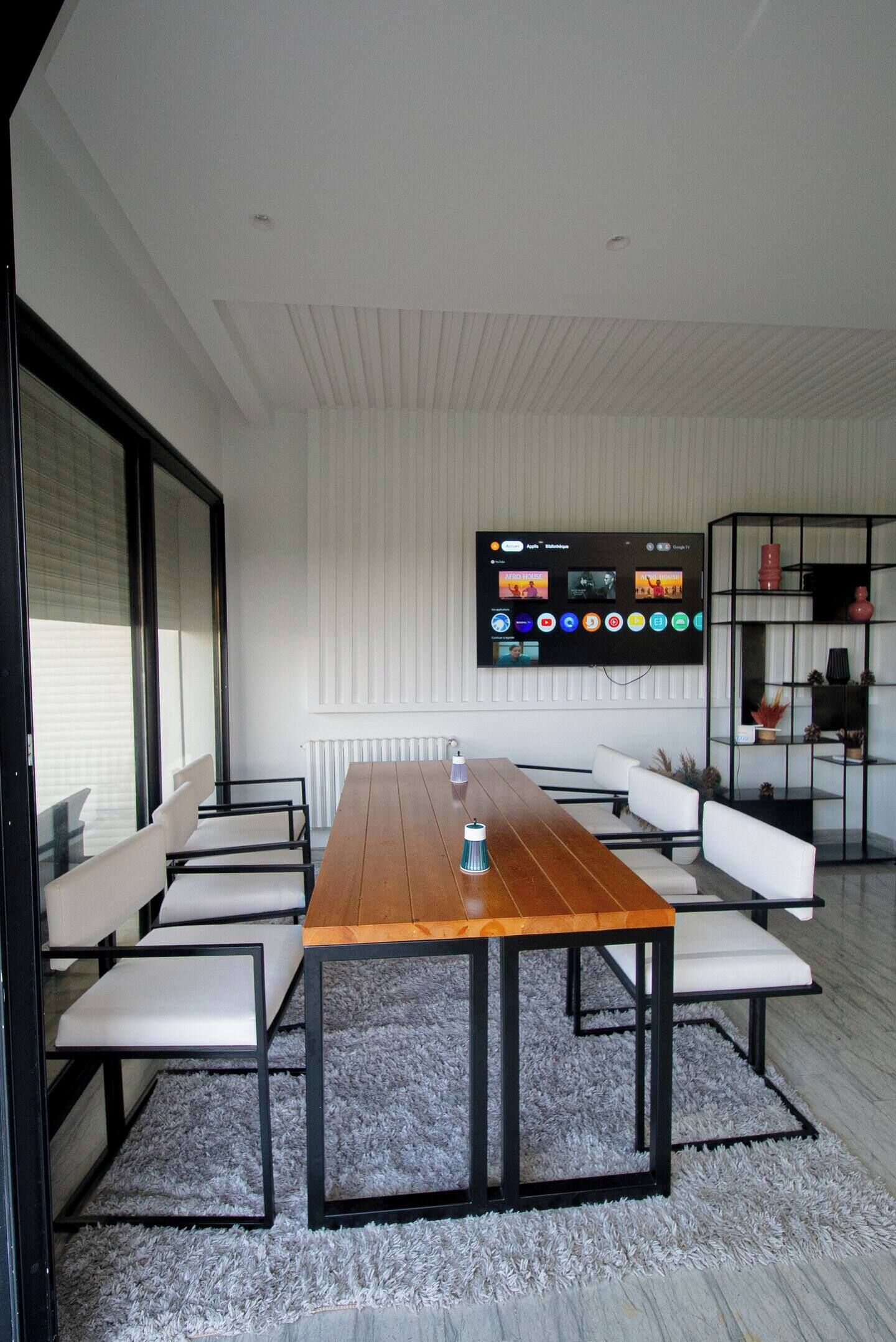 Un espace repas moderne avec une table en bois, des chaises blanches, un grand téléviseur à écran plat fixé au mur et une étagère noire contenant des objets décoratifs.
