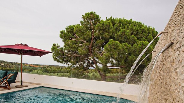 Una piscina amb cascada, envoltada d'una terrassa de rajola i un parasol vermell.
