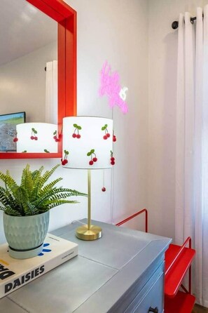Una habitación moderna con una lámpara blanca con detalles color cereza, un espejo de marco rojo, una planta en maceta y un libro titulado 'DAISIES'.