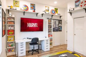 Una habitación con escritorio, silla, estanterías llenas de libros y juguetes, un televisor mostrando 'MARVEL' en la pantalla, y carteles de superhéroes.