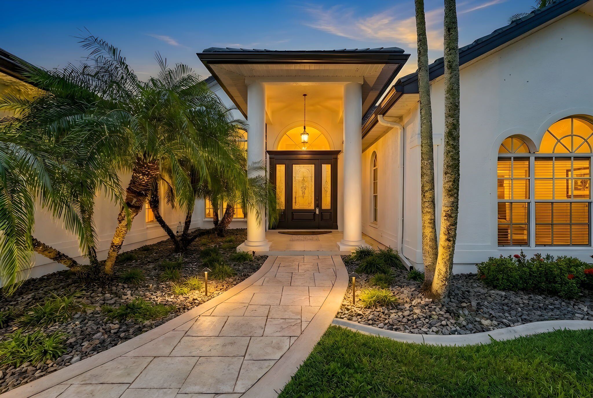 A well-lit pathway leading to a building with a covered entrance, palm trees, and a landscaped garden.