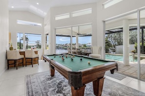 A pool table in a spacious room with large windows, a dining area, and a view of a pool.