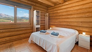 A wooden cabin bedroom with a bed, nightstands, a shelf unit, and a large window offering a mountain view. - Nordic cottage on the Costa del Sol, enjoy the Andalusian climate with swimming pool! (AN)