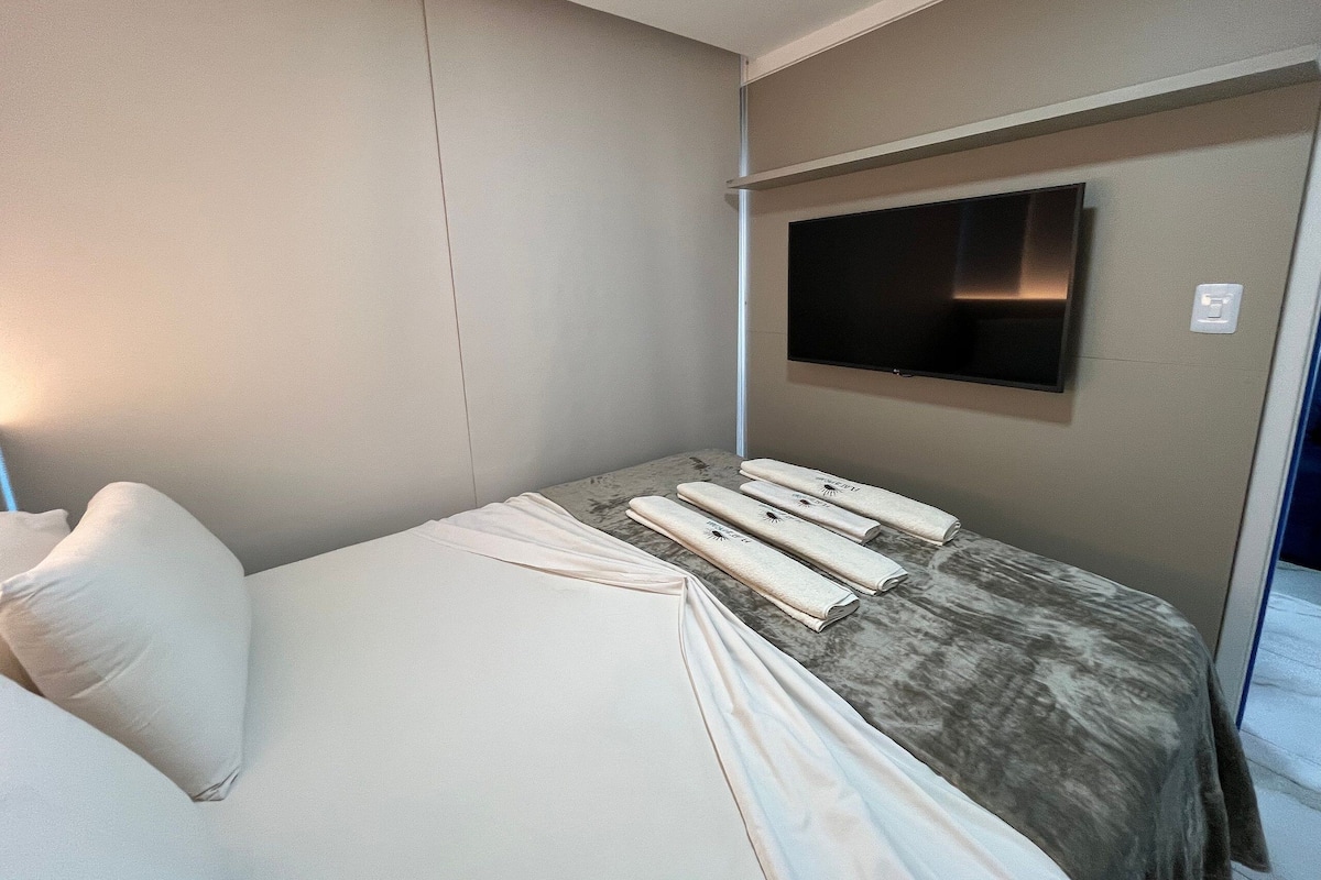 Moderni hotellihuone, jossa on sänky, seinälle asennettu taulutelevisio ja pieni pöytä, jonka päällä on pyyhkeitä.
