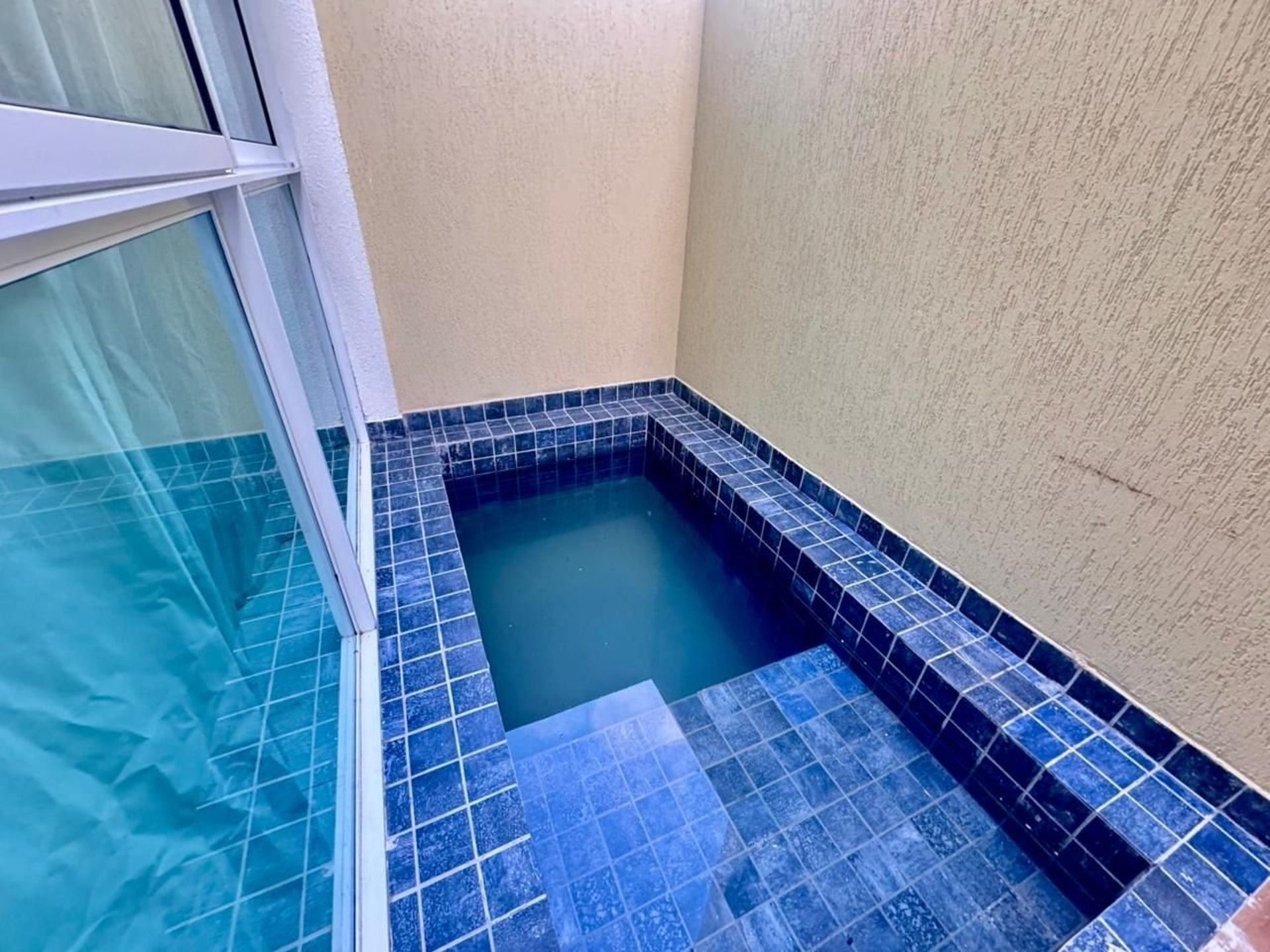 A small, square hot tub with blue tiles, surrounded by a beige wall and a glass window.