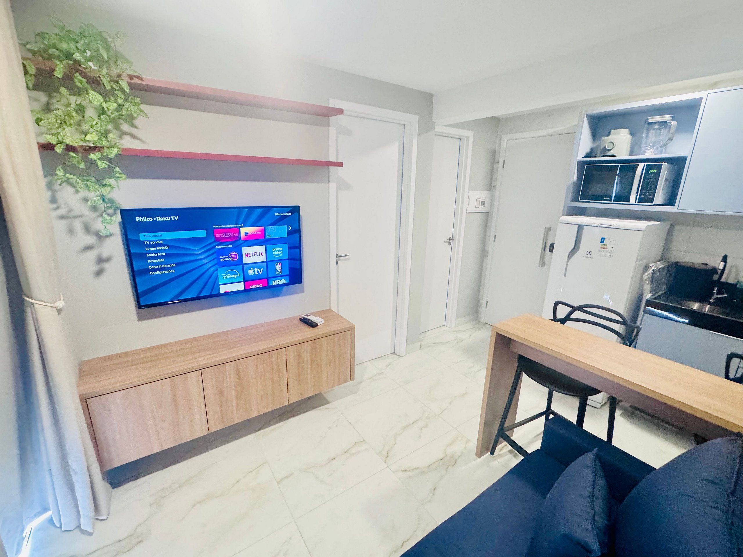 A modern living room with a flat-screen TV mounted on the wall, a wooden cabinet, a dining table with chairs, and a kitchen area with a microwave and refrigerator.